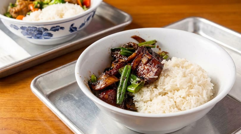 Two bowls of Asian cuisine with rice, meat, and vegetables on metal trays.