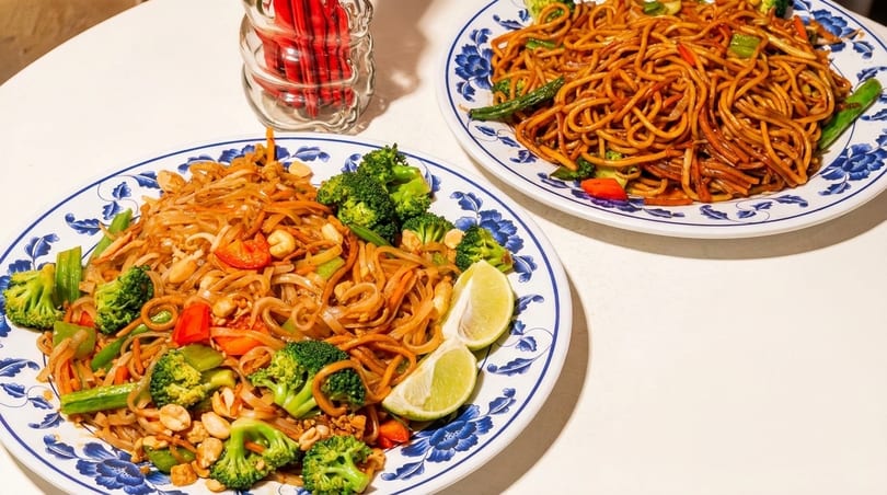 Two plates of noodles with broccoli, peppers, and peanuts, one with lime wedges.