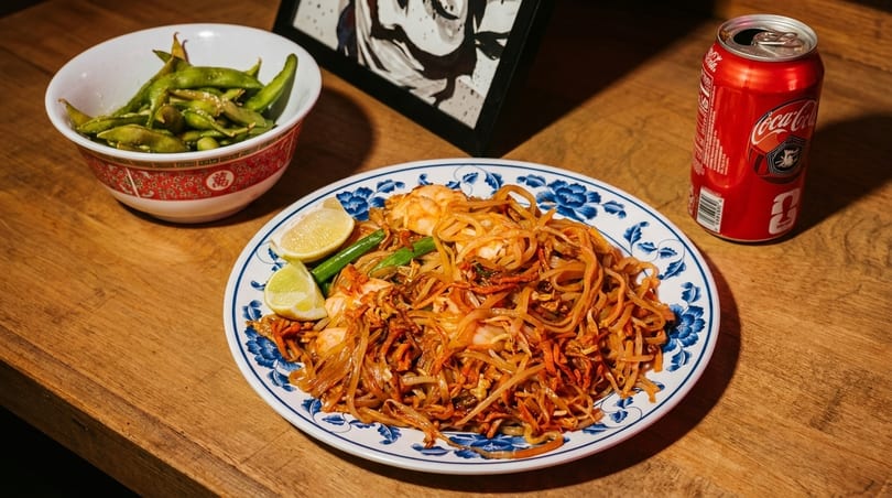 Pad Thai with shrimp and lime wedges, a bowl of edamame, and a Coca-Cola can on a wooden table.