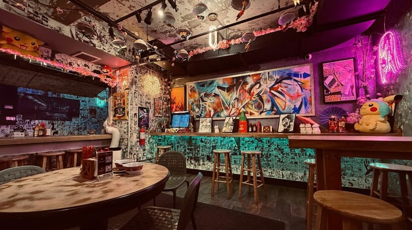A vibrant, anime-themed restaurant interior with manga-covered walls, neon lights, and colorful graffiti art.