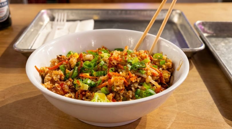 A white bowl of fried rice with carrots, green onions, and chopsticks on a wooden table.