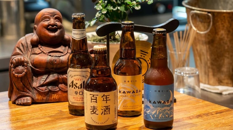 Four bottles of Japanese beer, including Asahi and Kawaba, on a wooden table next to a Buddha statue.